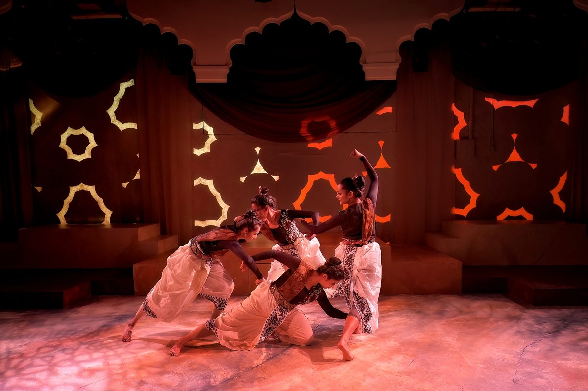 Five dancers in red skirts and gold-embroidered bodices mid-turn on a warmly lit stage, skirts flared wide. Behind them a painted set of pale stone arches, with more performers standing at the edges.